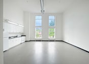 Thumbnail 19 of 22 - A large empty room with a kitchen area to the left and a window with a view of the outside.
