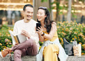 Thumbnail 10 of 22 - A man and woman are sitting on a bench with a book and a phone in their hands.