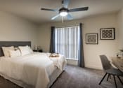 Thumbnail 22 of 47 - Staged bedroom in a 1-bedroom apartment at Cambridge Court Apartments in Dallas, Texas, featuring carpeted flooring, ceiling fan, patio door, desk area, and natural light.