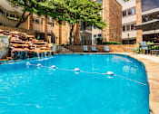 Thumbnail 31 of 42 - A swimming pool with a waterfall surrounded by lounge chairs and a couple of tables, chairs and umbrellas at Cambridge Court Apartments Entryway in Lake Highlands, Dallas, TX.
