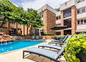 Thumbnail 33 of 42 - A swimming pool with a waterfall surrounded by lounge chairs and a couple of tables, chairs and umbrellas at Cambridge Court Apartments Entryway in Lake Highlands, Dallas, TX.