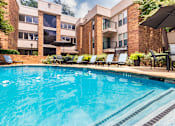 Thumbnail 32 of 42 - A swimming pool with a waterfall surrounded by lounge chairs and a couple of tables, chairs and umbrellas at Cambridge Court Apartments Entryway in Lake Highlands, Dallas, TX.