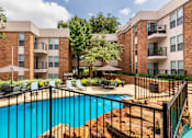 Thumbnail 30 of 42 - A swimming pool with a waterfall surrounded by lounge chairs and a couple of tables, chairs and umbrellas at Cambridge Court Apartments Entryway in Lake Highlands, Dallas, TX.