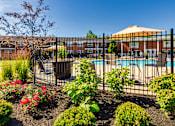 Thumbnail 50 of 59 - Pool entrance with black fencing, flowers, and umbrella-covered seating at Lake of the Woods Apartments