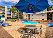 Thumbnail 46 of 62 - A pool area with lounge chairs, patio tables and chairs with blue umbrellas and stainless steel BBQ in front of apartment buildings at Princeton Court Apartments in the Vickery Midtown neighborhood of Dallas, TX.