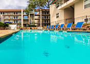 Thumbnail 12 of 62 - A pool area with lounge chairs, a patio table and chairs with a blue umbrella and stainless steel BBQ in front of apartment buildings at Princeton Court Apartments in the Vickery Midtown neighborhood of Dallas, TX.