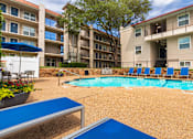 Thumbnail 13 of 62 - A pool area with lounge chairs, a patio table and chairs with a blue umbrella and stainless steel BBQ in front of apartment buildings at Princeton Court Apartments in the Vickery Midtown neighborhood of Dallas, TX.