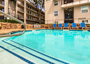 Thumbnail 45 of 62 - A pool area with lounge chairs, a patio table and chairs with a blue umbrella and stainless steel BBQ in front of apartment buildings at Princeton Court Apartments in the Vickery Midtown neighborhood of Dallas, TX.