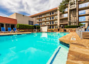 Thumbnail 47 of 62 - A pool area with lounge chairs, a patio table and chairs with a blue umbrella and stainless steel BBQ in front of apartment buildings at Princeton Court Apartments in the Vickery Midtown neighborhood of Dallas, TX.