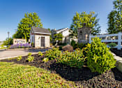 Thumbnail 90 of 96 - A small stone building with a black door is surrounded by a garden with a white fence in the background.