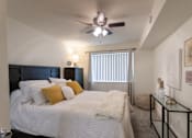 Thumbnail 15 of 44 - A bedroom with a bed, a ceiling fan, and a window with blinds in the 558 square foot 1 bedroom, 1 bath model apartment at The Summit at Midtown Apartments in the Dallas Midtown neighborhood of Dallas, TX.