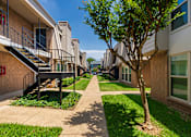 Thumbnail 27 of 31 - Walkway between apartment buildings with green showing landscaping at Woodbridge Apartments in Midtown, Dallas, TX.