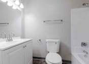 Thumbnail 15 of 66 - A white toilet with a blue sticker on the lid is in a bathroom with a white sink and a white cabinet.