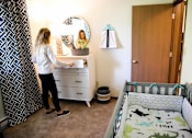 Thumbnail 13 of 90 - a woman standing in a nursery with a crib and a dresser at Rosemont Place Apartments, Des Moines, IA, 50320
