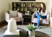 Thumbnail 2 of 26 - A woman is sitting on a chair in a living room with a TV mounted on the wall at Sutton Hill Apartments, Des Moines, IA