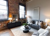 Thumbnail 31 of 99 - A living room with a grey couch, a black coffee table, and a fireplace with a brick wall.at Plant 1 Lofts, N. Newton Iowa   