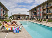Thumbnail 56 of 56 - A woman in a yellow bikini lounges by a pool with a man in a pink shirt at Meadowlark Place Apartments, Grimes, IA,  50111