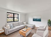 Thumbnail 11 of 49 - A living room with a large window, a white couch, a coffee table, and a painting on the wall.