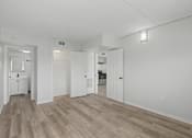 Thumbnail 12 of 26 - A white room with wood flooring and a kitchen area in the background.