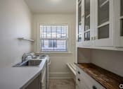 Thumbnail 26 of 27 - A kitchen with white cabinets and a window with blinds.