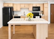 Thumbnail 33 of 35 - A kitchen with a black refrigerator, wooden cabinets, and a wooden island with a vase of yellow flowers on it.