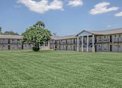 Thumbnail 9 of 12 - A large grassy field in front of a building with a tree in the middle.