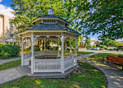 Thumbnail 4 of 18 - A gazebo with a black roof and white railings is surrounded by greenery.
