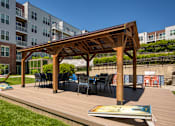 Thumbnail 14 of 35 - A wooden pergola with chairs and a table is in the foreground of a grassy area with apartment buildings in the background.
