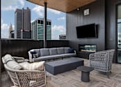 Thumbnail 26 of 55 - A modern balcony with a grey sofa, wicker chair, and a television.