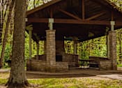 Thumbnail 55 of 61 - A pavilion with a stone base and a wooden roof is surrounded by trees.