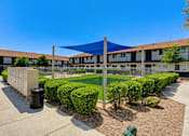 Thumbnail 9 of 28 - A sunny day at a residential complex with a blue shade structure and neatly trimmed bushes.
