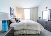 Thumbnail 8 of 15 - A bedroom with a large bed, a nightstand with a lamp, and a dresser with a mirror.