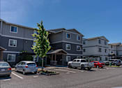 Thumbnail 10 of 22 - A parking lot with cars and apartment buildings in the background at St Helens Place Apartment Homes, St Helens, Oregon
