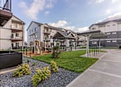 Thumbnail 6 of 49 - A residential area with apartment buildings and a small garden with bushes and a gravel path at Woodburn Place Apartment Homes, Oregon