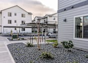 Thumbnail 5 of 49 - A white building with a grey roof is surrounded by a gravel area with plants at Woodburn Place Apartment Homes, Woodburn, OR, 97071