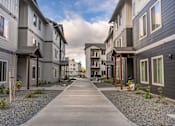 Thumbnail 2 of 49 - A long concrete walkway leads between two rows of modern townhouses at Woodburn Place Apartment Homes, Woodburn