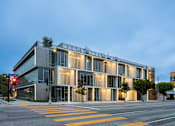 Thumbnail 1 of 9 - A modern building with a glass facade is illuminated from within and surrounded by a yellow crosswalk.