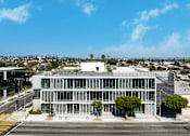 Thumbnail 3 of 9 - A modern building with a glass facade is situated on a street corner with trees and other buildings in the background.