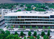Thumbnail 14 of 16 - A modern glass and steel building is surrounded by trees and parking.
