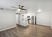 Thumbnail 8 of 23 - A room with a ceiling fan and a kitchen area in the background.