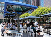 Thumbnail 5 of 18 - People are sitting at tables under blue umbrellas in an outdoor dining area.