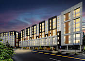 Thumbnail 36 of 38 - A modern building with a glass facade is illuminated from within, with a tree to the left and a street in front.