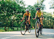 Thumbnail 3 of 11 - Two cyclists wearing helmets and sunglasses ride their bikes on a road.