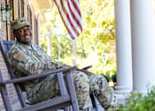 Thumbnail 36 of 74 - A soldier in uniform sits on a porch with an American flag in the background.