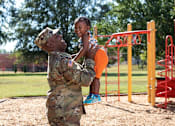 Thumbnail 5 of 37 - A soldier in camouflage uniform is lifting a young girl in a playground.