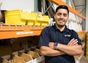 Thumbnail 8 of 37 - A man in a black shirt with his arms crossed in front of a shelf with boxes and containers.