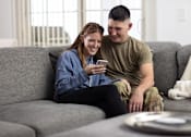 Thumbnail 11 of 37 - A man and a woman are sitting on a grey couch, with the woman looking at her phone.