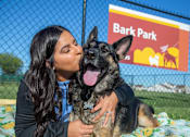 Thumbnail 4 of 21 - A woman is hugging a German Shepherd dog in front of a sign that says Bark Park.