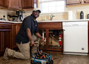 Thumbnail 1 of 21 - A man in a black shirt and khaki pants is kneeling on the floor in front of a washing machine.
