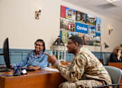 Thumbnail 6 of 21 - A man in military uniform is talking to a woman in a denim jacket in an office setting.